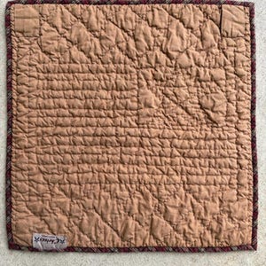 May include: A square, quilted placemat in a warm brown hue. The surface features a diamond pattern, with a plaid border in burgundy and brown. The label reads "Choices".