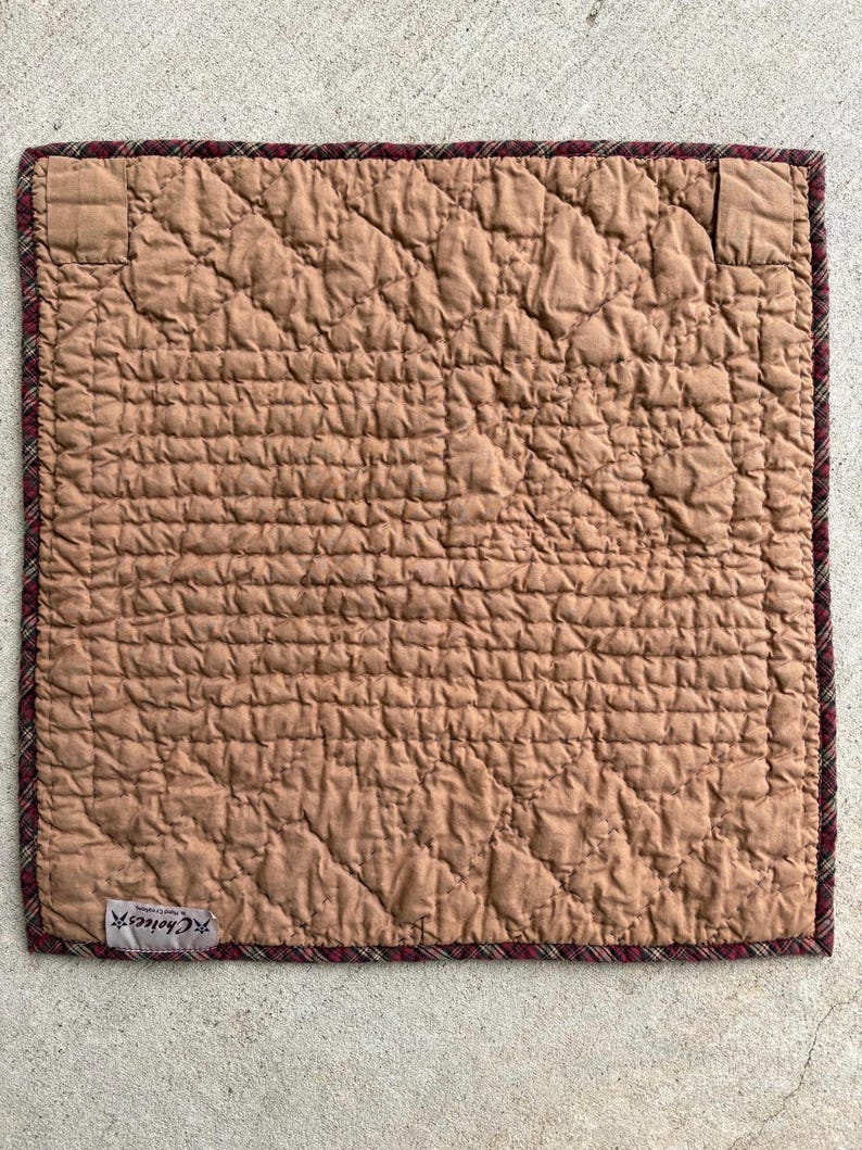 May include: A square, quilted placemat in a warm brown hue. The surface features a diamond pattern, with a plaid border in burgundy and brown. The label reads "Choices".