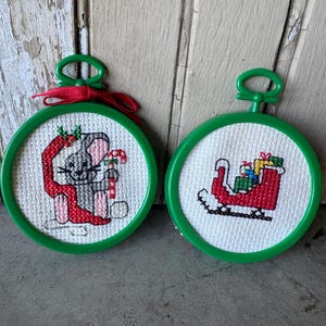 May include: Two Christmas ornaments with green frames and white cross-stitch designs. One features a gray mouse with a candy cane and red ribbon. The other shows a red sleigh filled with gifts. A red ribbon is attached to the first ornament.