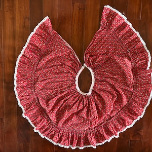 May include: A red skirt featuring a small floral pattern in white and green. The garment has a wide, circular silhouette with a gathered, ruffled hemline trimmed with white lace along the entire outer edge and the central waist opening.
