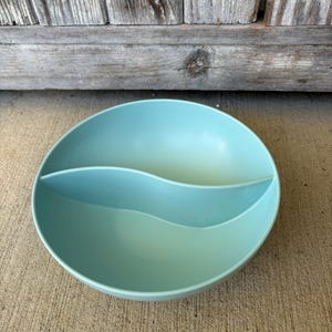 May include: A round, light blue serving bowl with a wave-shaped divider. The bowl is made of a solid material and has a smooth, matte finish. The bowl is designed to hold two different types of food.
