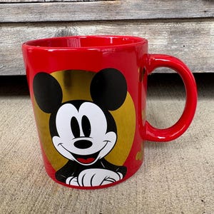 May include: A red ceramic mug featuring Mickey Mouse. The mug has a gold background behind Mickey's face and black ears. The handle is red, and the mug is sitting on a concrete surface.