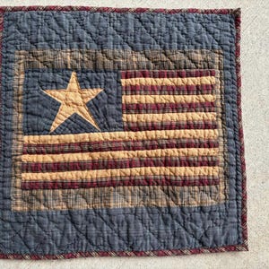 May include: A quilted square featuring an American flag design. The flag has a blue field with a tan star, and alternating tan and burgundy stripes. The border is a dark blue with a burgundy trim. The quilt is made of fabric.