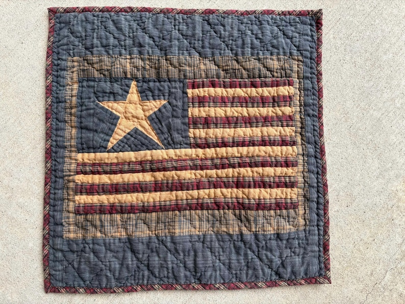 May include: A quilted square featuring an American flag design. The flag has a blue field with a tan star, and alternating tan and burgundy stripes. The border is a dark blue with a burgundy trim. The quilt is made of fabric.