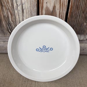 May include: White ceramic pie plate with a blue floral design in the center. The plate has a wide rim and is set against a wooden background. A classic kitchenware item.