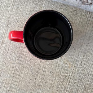May include: A red coffee mug with a black interior, viewed from above. The mug has a rounded shape with a handle on the side. The mug is sitting on a concrete surface.