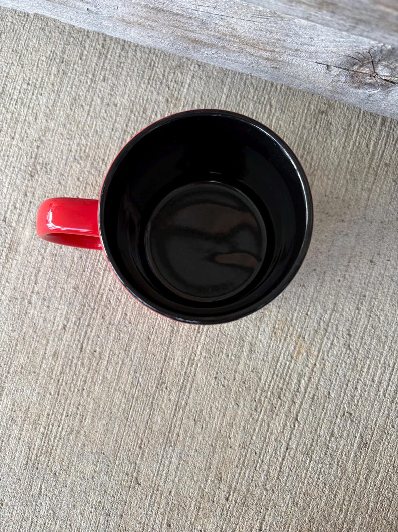 May include: A red coffee mug with a black interior, viewed from above. The mug has a rounded shape with a handle on the side. The mug is sitting on a concrete surface.