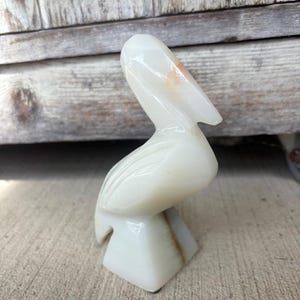 May include: A carved white onyx pelican sculpture. The bird is depicted with its head tilted back, and its beak is slightly open. The sculpture has a smooth, polished finish and sits on a square base. The onyx has subtle brown veining.