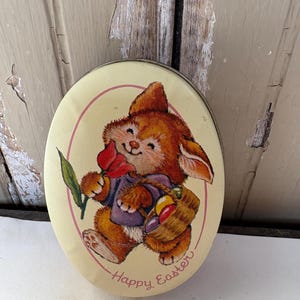May include: Oval-shaped Easter tin with a cartoon bunny illustration. The bunny is brown and wearing a purple shirt, holding a red flower and a basket of Easter eggs. The tin has a light yellow background with the words "Happy Easter".