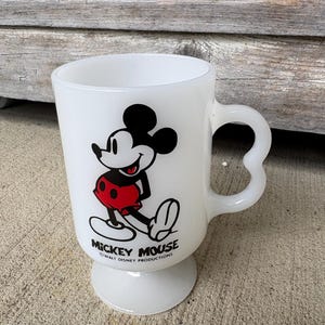 Może przedstawiać: Biały kubek z mlecznego szkła na n&oacute;żce z grafiką Myszki Miki w kolorach czarnym, białym i czerwonym. Pod postacią nadrukowano napis MICKEY MOUSE oraz WALT DISNEY PRODUCTIONS. Kubek posiada wygięte ucho oraz okrągłą podstawę.