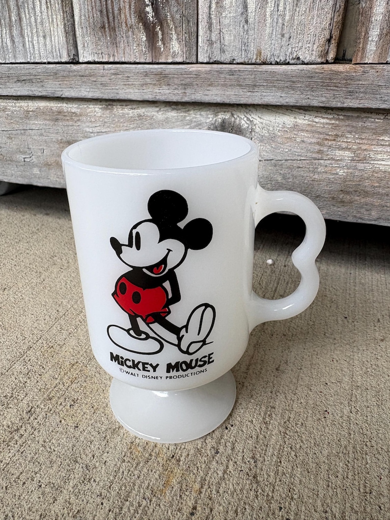Może przedstawiać: Biały kubek z mlecznego szkła na n&oacute;żce z grafiką Myszki Miki w kolorach czarnym, białym i czerwonym. Pod postacią nadrukowano napis MICKEY MOUSE oraz WALT DISNEY PRODUCTIONS. Kubek posiada wygięte ucho oraz okrągłą podstawę.