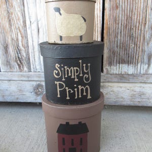 Puede incluir: Tres cajas decorativas redondas apiladas. La caja superior es de color canela con una ilustración de oveja. La caja del medio es negra con las palabras "Simply Prim" en crema. La caja inferior es marrón con una ilustración de casa roja.