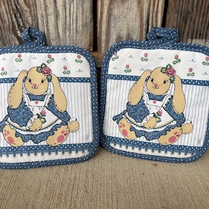 May include: Two square pot holders with a bunny design. The bunnies are tan with pink accents and wear blue dresses. The pot holders have a white background with vertical stripes and a blue border with a loop for hanging.