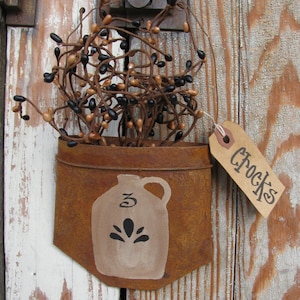 May include: A rustic, metal pocket with a painted jug design and the word "Crocks" on a tag. The pocket holds a bundle of brown and black berry stems, hanging on a weathered wooden door.