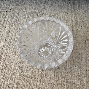 May include: Clear glass ashtray with a circular shape and a detailed, starburst pattern. The ashtray has a scalloped edge and a central well. The glass is transparent, allowing light to pass through, and is sitting on a concrete surface.