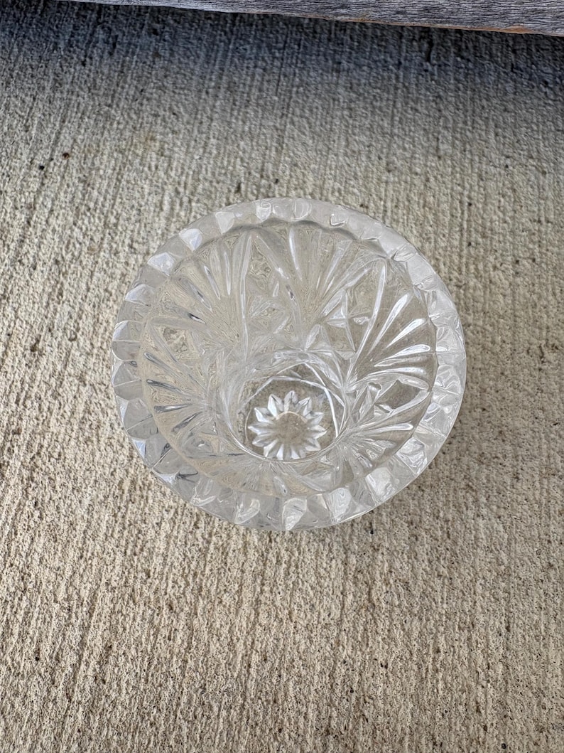 May include: Clear glass ashtray with a circular shape and a detailed, starburst pattern. The ashtray has a scalloped edge and a central well. The glass is transparent, allowing light to pass through, and is sitting on a concrete surface.