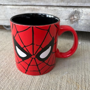 May include: A red ceramic mug featuring a Spider-Man design. The mug has a black interior and a red handle. The design includes a black web pattern and white eye shapes, resembling Spider-Man's mask.