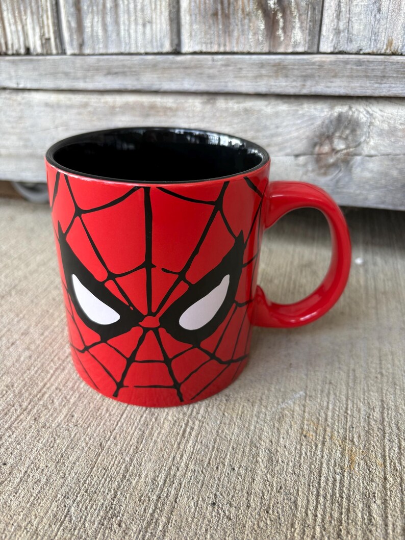 May include: A red ceramic mug featuring a Spider-Man design. The mug has a black interior and a red handle. The design includes a black web pattern and white eye shapes, resembling Spider-Man's mask.