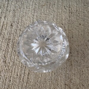 May include: A clear, cut-glass ashtray with a starburst pattern on the bottom. The ashtray is round with a scalloped edge. The glass is transparent, allowing light to pass through. The ashtray is sitting on a concrete surface.