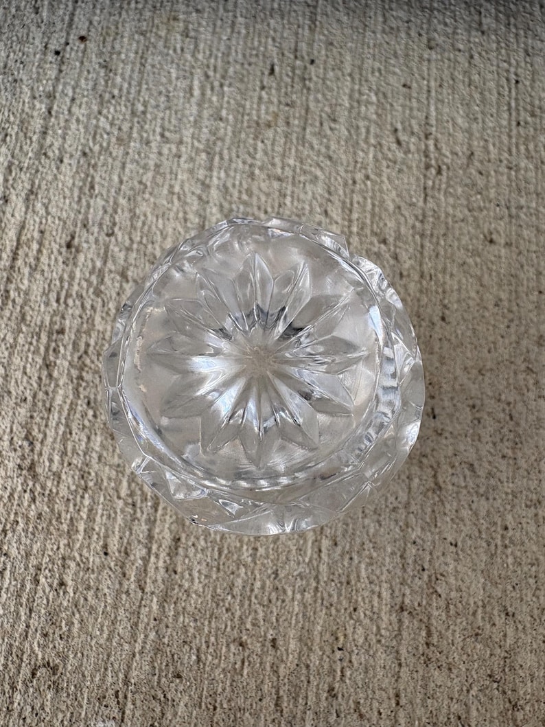 May include: A clear, cut-glass ashtray with a starburst pattern on the bottom. The ashtray is round with a scalloped edge. The glass is transparent, allowing light to pass through. The ashtray is sitting on a concrete surface.