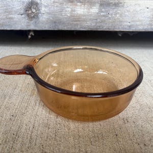 May include: Amber-colored glass measuring cup with a handle. The bowl is round with a dark brown rim. The handle is oval-shaped and attached to the side of the bowl. The cup is sitting on a concrete surface.