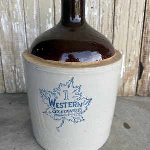 May include: A vintage stoneware jug with a brown glazed top and a light beige base. The jug features a blue maple leaf design with the text "1 Western Stoneware Co. Monmouth Ill." and a cork stopper.