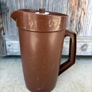 May include: A brown plastic pitcher featuring a cylindrical body, a sturdy side handle, and a matching lid with a small central knob. The pitcher has a molded pouring spout and a textured surface finish, showing minor signs of surface wear.