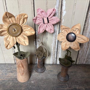 May include: Three handmade fabric flowers in shades of tan and pink. One flower is in a small vase, another is on a wooden spool, and the third is also on a wooden spool. The flowers have button centers and are arranged against a rustic wooden background.