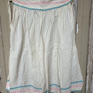 May include: An off-white apron featuring a gathered waist with tie closures. The waistband and hem are decorated with horizontal rows of teal and pink rickrack trim. The fabric has a natural, textured appearance with vertical folds throughout.