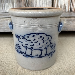 May include: A cylindrical stoneware crock with a light gray speckled finish and two blue-glazed side handles. The front features a blue hand-painted sheep graphic and a stamped logo reading "Iowa Pottery Works" with the year 1988 below the text.