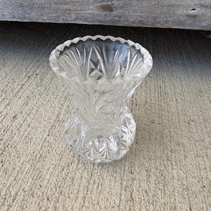 May include: A clear, cut-glass vase with a flared top and a rounded base. The vase features a detailed, geometric pattern throughout its design. The rim has a scalloped edge. The vase is sitting on a concrete surface.