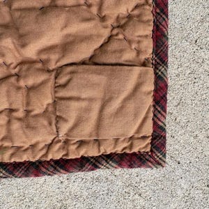 May include: Close-up of a quilted blanket with a brown center and a plaid border. The blanket has a stitched pattern and a rectangular pocket. The border features a red, black, and green plaid design.