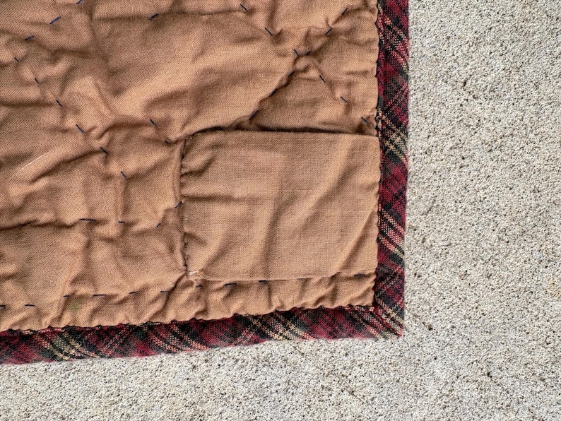 May include: Close-up of a quilted blanket with a brown center and a plaid border. The blanket has a stitched pattern and a rectangular pocket. The border features a red, black, and green plaid design.