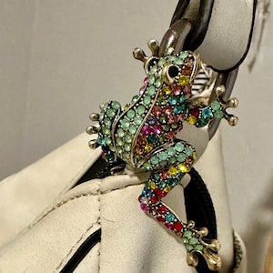 May include: A jeweled frog-shaped accessory with a variety of colorful rhinestones, including green, pink, and yellow. The frog is attached to a black cord and hangs from a white handbag.