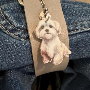 May include: A gray leather keychain with a silver metal clasp and a charm featuring a white dog with brown eyes.