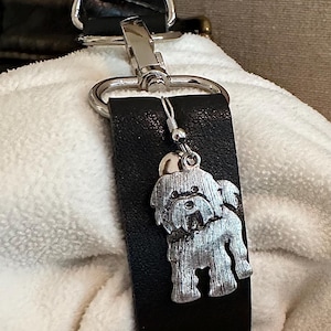 May include: A black leather bag charm with a silver dog charm attached. The dog charm is a detailed depiction of a small, fluffy dog.