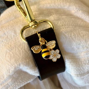 May include: A gold-colored keychain clip attached to a black leather strap. A charm with a bee design, featuring yellow and black stripes, is adorned with rhinestones on the wings. A white flower charm is also attached.