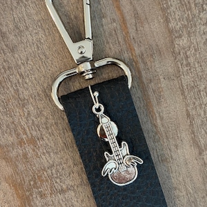 May include: A black leather strap with a silver-colored clasp and a guitar charm. The guitar charm has wings and is attached to the strap with a small silver hook. The background is a light brown wooden surface.