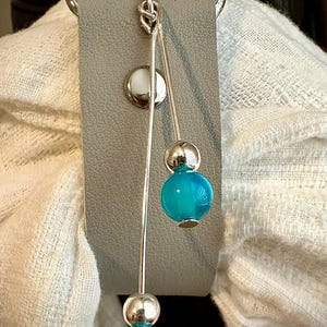 May include: A silver charm with a blue glass bead and a silver bead. The charm is attached to a gray leather strap.