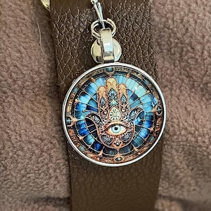 May include: A round pendant with a Hamsa hand design, featuring blue and gold details, hangs from a silver-colored clasp. The pendant is attached to a brown leather strap, with a silver-colored metal loop at the top.
