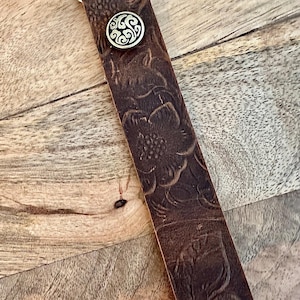 May include: Close-up of a brown leather strap with a floral embossed design. The strap features a silver-colored metal clasp and a decorative button with a swirling pattern. The leather has a distressed appearance.