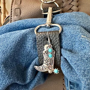 May include: Silver-toned cowboy boot charm with turquoise-colored accents. The charm is attached to a black leather strap, hanging from a silver clasp. The background includes a denim fabric and a brown leather bag.