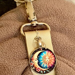 May include: A gold-colored keychain with a silver clasp and a round pendant. The pendant features a celestial design with a crescent moon and a sun, in shades of blue, red, and yellow. The keychain is attached to a tan strap.