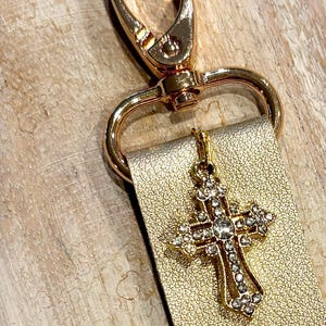 May include: Gold-toned keychain with a decorative cross pendant. The cross is encrusted with clear crystals. The keychain features a gold clasp and a gold leather strap. The background is a light brown wooden surface.
