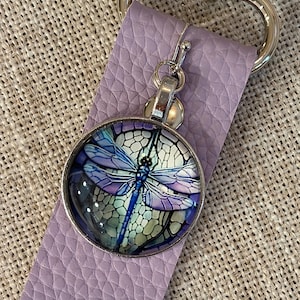 May include: A round, silver-toned keychain charm featuring a dragonfly design in shades of blue, purple, and green. The charm is attached to a lavender leather strap with a silver clasp. The dragonfly design is detailed and artistic.