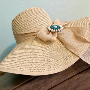 May include: A beige straw sun hat with a wide brim and a textured weave. The hat features a large beige fabric bow wrapped around the crown, accented by a circular brooch with a central turquoise stone surrounded by white oval beads.