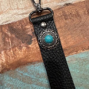 May include: Black leather wrist strap with a silver-tone clasp and a decorative turquoise-colored stone set in a silver-tone filigree. The strap has a textured surface and a metal rivet.