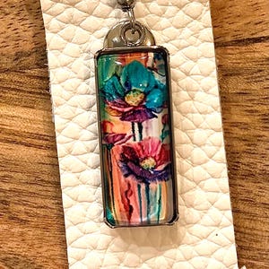 May include: A rectangular earring with a floral design. The image features a close-up of a silver-toned earring with a colorful floral print. The flowers are in shades of teal, pink, and purple, set against a white background.