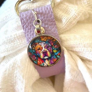 May include: A dog-themed pendant with a colorful, stained-glass style design. The pendant is attached to a lavender leather strap and a silver-colored clasp. The dog's portrait features vibrant hues of orange, blue, purple, and yellow.
