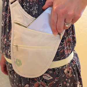 May include: A light beige crossbody bag with multiple zippered compartments. The bag has a small, embroidered flower detail and a long, adjustable strap. A smartphone is partially visible in the front pocket.